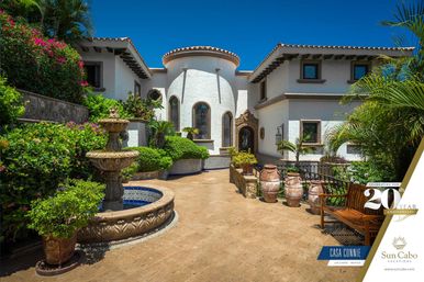 Sun-drenched Los Cabos villa courtyard with white stucco Mediterranean-style architecture, ornate stone fountain, terracotta urns, lush tropical plants and bright blue sky.