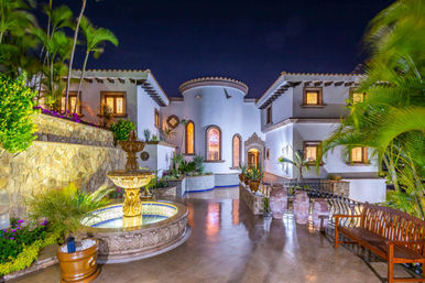 Night view of a Mediterranean-style villa courtyard with a glowing tiered fountain, arched windows, tiled accents, stone walls, palm trees, potted planters and a polished stone patio.