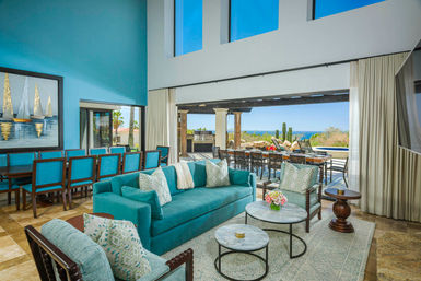 Sunlit coastal living room in a beachfront villa with a turquoise sofa and patterned pillows, matching dining chairs, tall windows and sliding doors opening to a shaded patio with outdoor dining, grill and ocean views.
