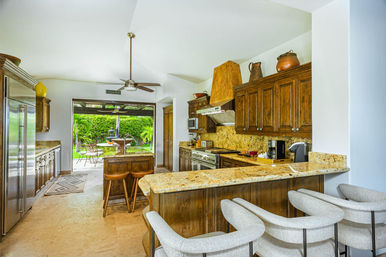 Sunlit open-concept kitchen with granite countertops and warm wood cabinets, island with wooden stools, bar seating with upholstered chairs, stainless steel appliances, ceiling fan, and sliding doors to a patio overlooking a lush garden and fountain.