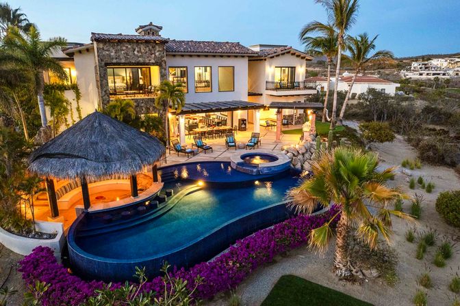 Aerial dusk view of a luxury coastal villa with a curved infinity pool, thatched palapa bar, raised spa, outdoor lounge seating, palm trees and bright bougainvillea set against a sandy coastal landscape.
