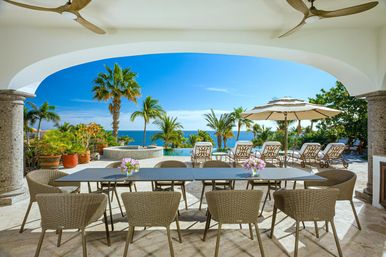Sunny tropical oceanfront terrace with wicker dining table and chairs, infinity pool, palm trees, lounge chairs and umbrella overlooking bright blue sea.