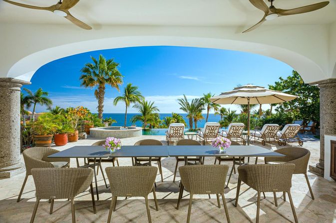 Sunny tropical oceanfront terrace with wicker dining table and chairs, infinity pool, palm trees, lounge chairs and umbrella overlooking bright blue sea.