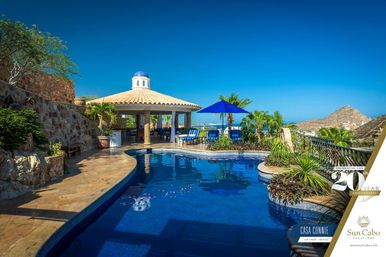 Luxury ocean-view pool terrace at a seaside villa in Los Cabos, Mexico — blue-tiled freeform pool, shaded domed cabana, palm trees and loungers under a bright blue sky.