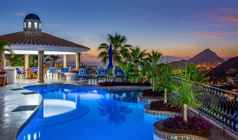 Ocean-view resort terrace at twilight — curved blue pool with tiled planters and palm trees, lounge seating under a domed pavilion and distant mountain silhouette.