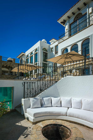 Sunlit Mediterranean-style coastal villa terrace with white stucco walls, arched windows, wrought-iron railings, beige patio umbrellas and a curved white sectional sofa around a stone fire pit beneath a clear blue sky.