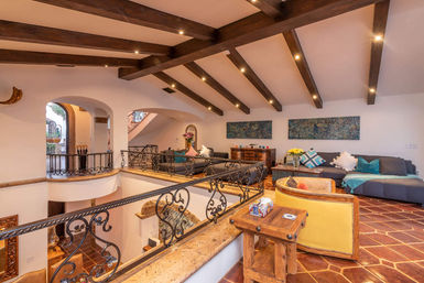 Sunlit Mediterranean-style loft living room with dark exposed wood beams, decorative wrought-iron balcony railing overlooking a lower level, terracotta tile floors, cozy sofas with colorful pillows, wooden tables and fresh flowers.