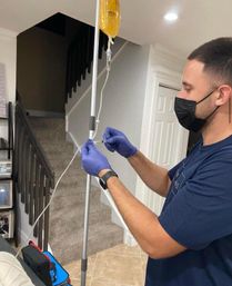 Masked caregiver in purple gloves adjusting a yellow hydration IV bag and tubing on a pole inside a home near a carpeted staircase
