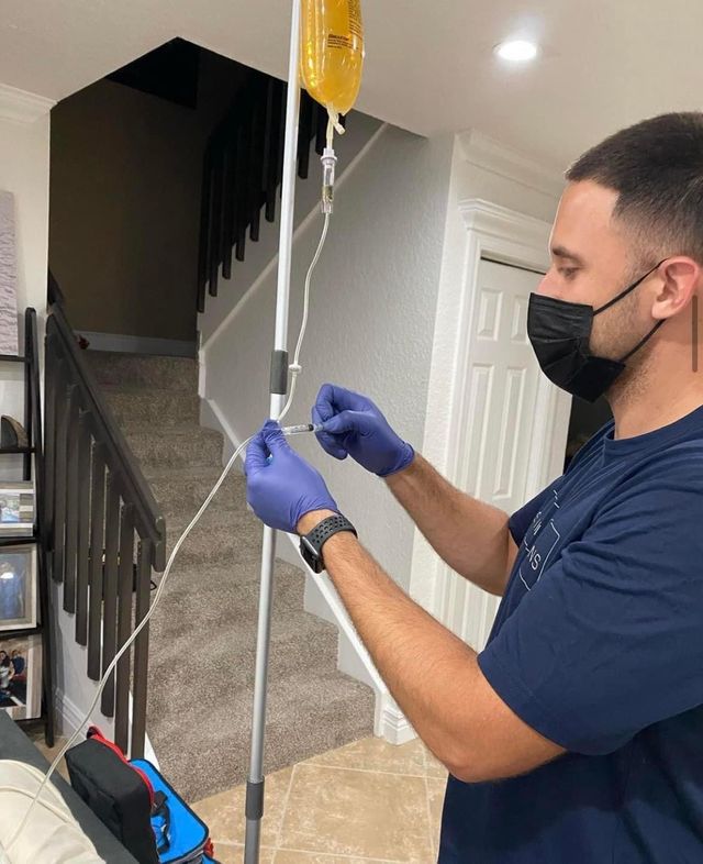 Medical professional in black mask and purple gloves primes tubing with a syringe from a yellow IV bag on a pole in a home hallway next to carpeted stairs — mobile IV therapy.