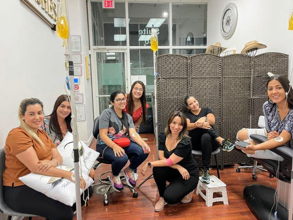 Group of seven smiling women relaxing while receiving IV drips in a bright wellness clinic lounge.