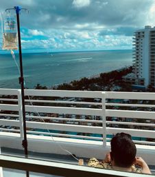 IV drip bag on a pole at an oceanfront hotel balcony overlooking turquoise sea, sandy beach, palm-lined resort pools and a high-rise, with a person seated facing the view