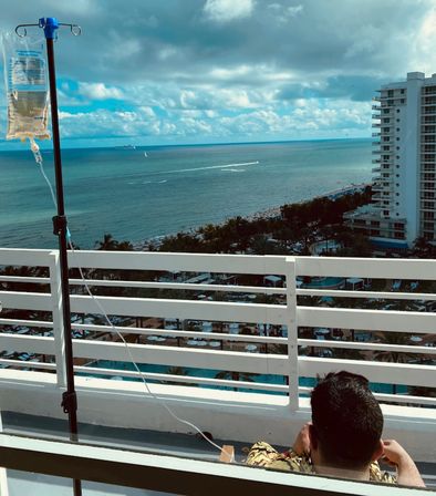 IV drip bag on a pole at an oceanfront hotel balcony overlooking turquoise sea, sandy beach, palm-lined resort pools and a high-rise, with a person seated facing the view