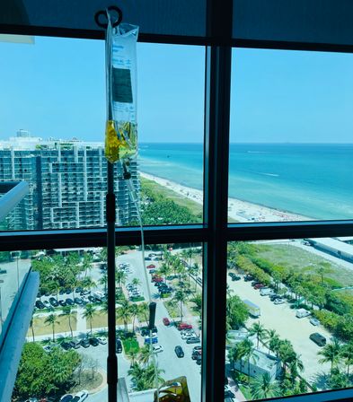 IV drip bag with yellow fluid hanging by a high-rise window overlooking a coastal city scene with palm trees, parking lot, sunlit turquoise ocean and sandy beach.
