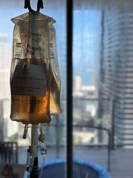 IV infusion bag of amber fluid hanging by a sunlit high-rise window, blurred downtown skyscrapers and balcony trampoline in the background.