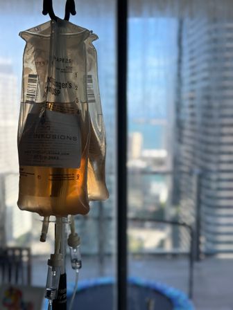 IV infusion bag of amber fluid hanging by a sunlit high-rise window, blurred downtown skyscrapers and balcony trampoline in the background.