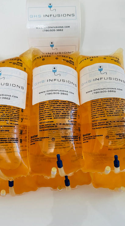 Bright amber IV infusion bags with clear medical labels and blue ports arranged on a white surface — intravenous vitamin drip and hydration therapy supplies.