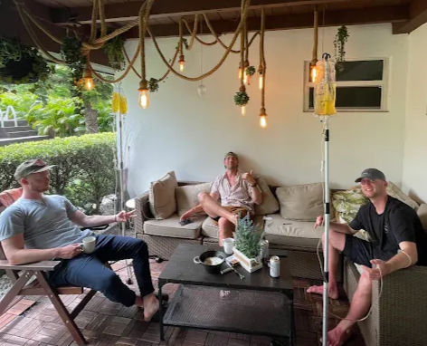 Three men relaxing on a covered backyard patio lounge with wicker sofa and chairs, hanging rope Edison bulbs, coffee mug and beer can on a low table, potted centerpiece and an IV drip pole with yellow fluid, with lush tropical greenery outside.