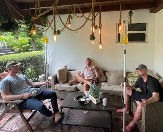 Three men relaxing on a covered backyard patio lounge with wicker sofa and chairs, hanging rope Edison bulbs, coffee mug and beer can on a low table, potted centerpiece and an IV drip pole with yellow fluid, with lush tropical greenery outside.