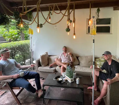 Three men relaxing on a covered backyard patio lounge with wicker sofa and chairs, hanging rope Edison bulbs, coffee mug and beer can on a low table, potted centerpiece and an IV drip pole with yellow fluid, with lush tropical greenery outside.