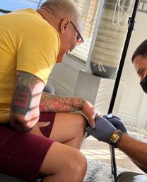 Person in a yellow shirt and burgundy shorts with colorful arm tattoos and blue nail polish receiving in-home IV care from a gloved, masked provider beside an IV pole in a sunlit living room.