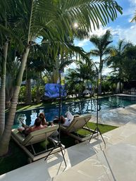 Tropical backyard pool scene with two people and a dog relaxing on chaise lounges under palm trees, outdoor TV, IV-hydration stands, and sunny blue sky