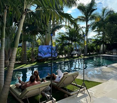 Tropical backyard pool scene with two people and a dog relaxing on chaise lounges under palm trees, outdoor TV, IV-hydration stands, and sunny blue sky