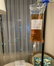 Amber IV drip bag hanging from an IV pole in a hotel room next to sheer curtains, a chair and a small table with medical supplies — intravenous therapy setup.