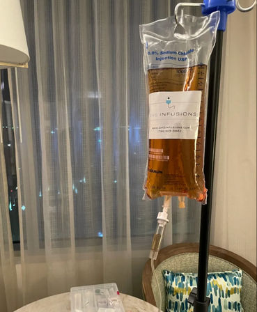 Amber IV drip bag hanging from an IV pole in a hotel room next to sheer curtains, a chair and a small table with medical supplies — intravenous therapy setup.