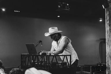 Black-and-white photo of a cowboy-hat DJ in a sleeveless shirt and sunglasses leaning over a laptop and mixer on a small live-music venue stage with a microphone and drinks nearby.