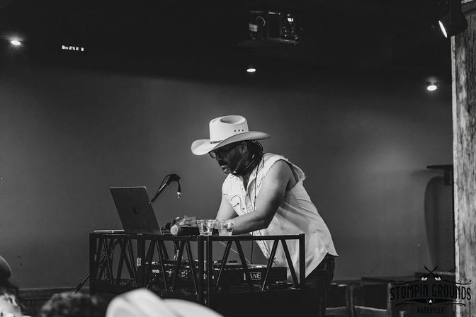 Black-and-white photo of a cowboy-hat DJ in a sleeveless shirt and sunglasses leaning over a laptop and mixer on a small live-music venue stage with a microphone and drinks nearby.