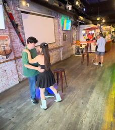 Two couples dancing on a small wooden dance floor inside a rustic bar with exposed brick walls, stools and colorful lights — woman in a black dress and white boots dancing with her partner.