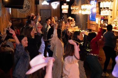 Energetic crowd line-dancing in a dim wooden bar or saloon, raised hands and a cowboy hat in the foreground with warm hanging lights over the bar.