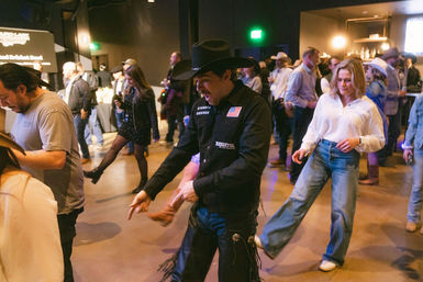 Crowd line dancing at an indoor country-western event — man in a black cowboy hat and chaps leading steps while a woman in a white blouse and wide-leg jeans follows, bar and other dancers in the background.