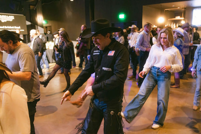 Crowd line dancing at an indoor country-western event — man in a black cowboy hat and chaps leading steps while a woman in a white blouse and wide-leg jeans follows, bar and other dancers in the background.
