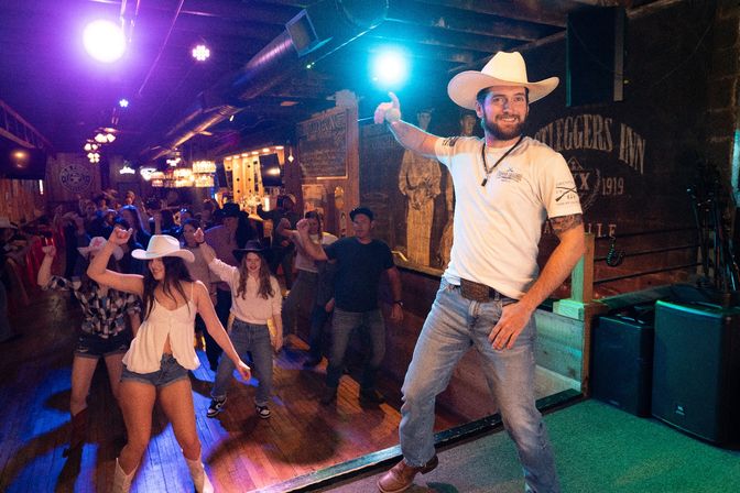 Man in a cowboy hat leading a lively line dance at a crowded country bar dance floor under colorful stage lights.