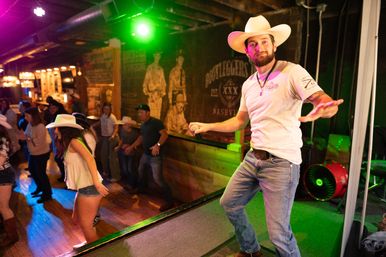 Male performer in a cowboy hat on stage holding a microphone, leading a lively line-dancing crowd in a neon-lit country bar/honky-tonk