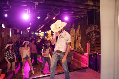 Male singer in a cowboy hat and jeans performing with a microphone on a small stage while a crowd of people in cowboy hats and boots line-dance under purple club lights in a rustic country bar.