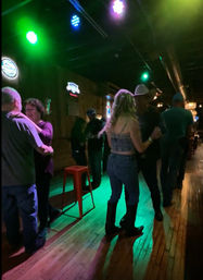Couple dancing on wooden floor in a dim country-style bar, woman in denim and boots and man in a cowboy hat, colorful stage lights and neon signs overhead