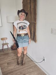 Smiling woman in a cowgirl hat wearing a graphic motorcycle crop tee, denim mini skirt, ornate belt and cowboy boots, holding a folded black umbrella in a boho‑chic entryway with macramé wall hanging and wood floors.