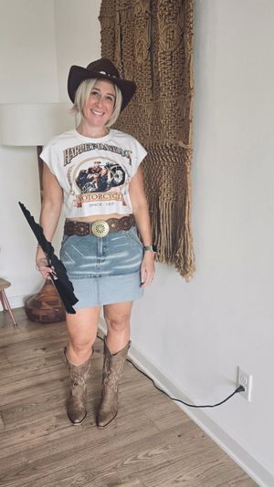 Smiling woman in a cowgirl hat wearing a graphic motorcycle crop tee, denim mini skirt, ornate belt and cowboy boots, holding a folded black umbrella in a boho‑chic entryway with macramé wall hanging and wood floors.