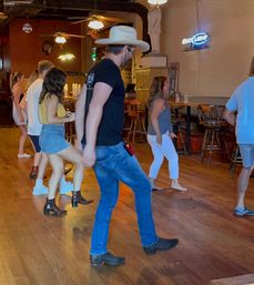 Line dancers at a rustic country bar on a wooden floor, instructor in a cowboy hat and jeans leading a mixed-age group in boots and casual summer clothes.