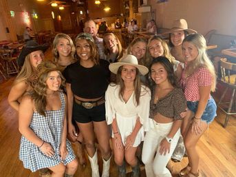 Smiling group of friends at a western-style bar, women wearing cowboy hats, boots and casual summer outfits, wooden floors and bar tables in the background — lively country night out.