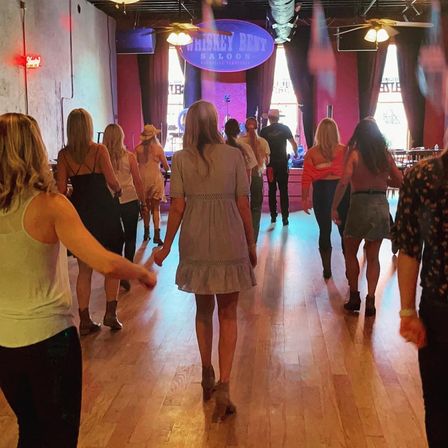 Group line dancing in a rustic country saloon with wooden dance floor, stage and warm ambient lighting