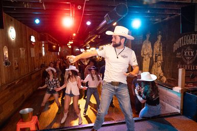 Energetic country singer in a cowboy hat on a small wooden bar stage passing the microphone as a crowd in boots and cowboy hats line-dances under colorful lights — Nashville country bar vibe