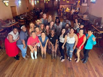 Group of about 20 smiling adults posing inside a rustic country-style bar or dance hall with wooden floors, neon beer signs, tables and booths; casual outfits, cowboy boots and hats, lively night-out vibe.