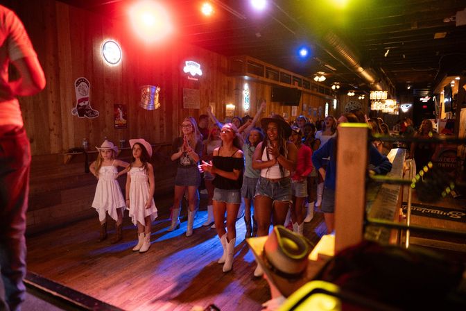 Group of girls and women line-dancing at a rustic country dance hall with colorful stage lights, cowboy hats and white boots, lively crowd near the stage