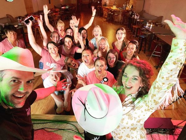 Stage selfie at a country-style bar: two hosts in cowboy hats pose with a cheering group of women on a wooden dance floor, tables and bar in the background — lively celebration vibe.