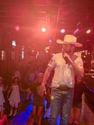 Male singer in a cowboy hat and white T‑shirt performs on a country bar stage, holding a microphone as a crowd of boot‑wearing dancers line up on a wooden dance floor under red stage lights.