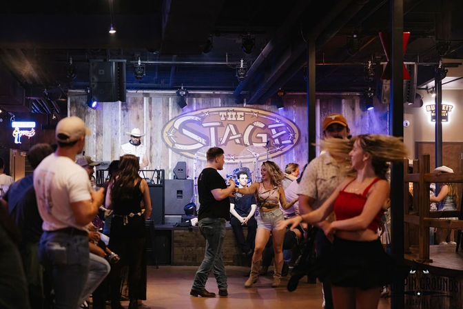 Lively indoor country dance hall with couples and friends dancing on the floor, wooden stage with live DJ in a cowboy hat, denim and boots, upbeat nightlife vibe