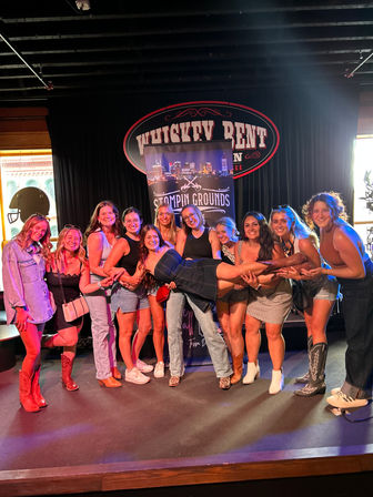 Group of friends posing on a downtown live-music stage at a country venue, one woman held horizontally while others smile, many wearing cowboy boots and colorful stage lighting
